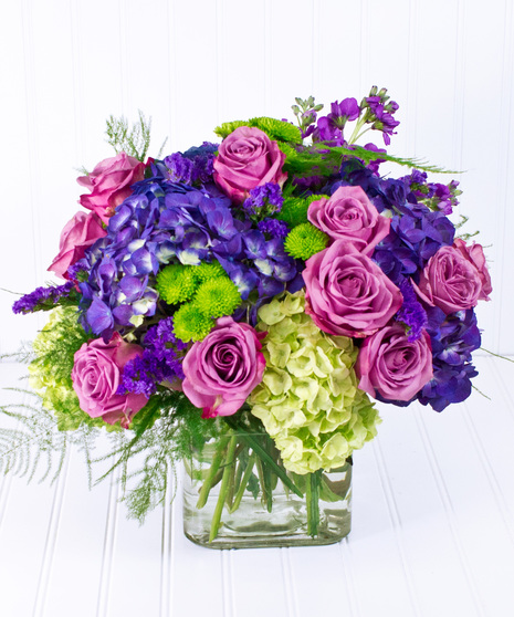 A glass cube vase, filled with lavender roses, and hydrangea accented with bright purple status.