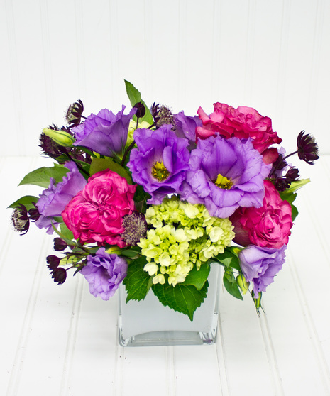 A glass cube vase, filled with lavender roses, and hydrangea accented with bright purple status.