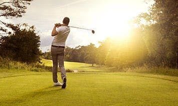 A golfer in mid-swing plays on a lush, green golf course surrounded by trees during a bright, sunny day.