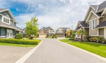 A curved residential street features several modern houses with well-maintained lawns and driveways under a partly cloudy sky, creating a peaceful suburban neighborhood scene.