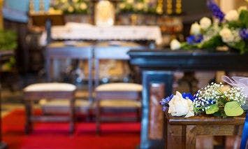 A small bouquet of flowers sits on a wooden pew in a church, which is decorated with an altar in the background and a red carpet.